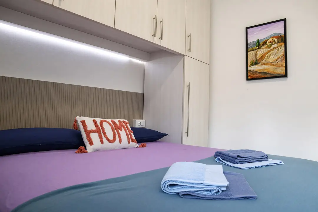 La camera dell’appartamento Lavanda: copriletto lilla e cuscino “HOME” sotto l’armadio a ponte