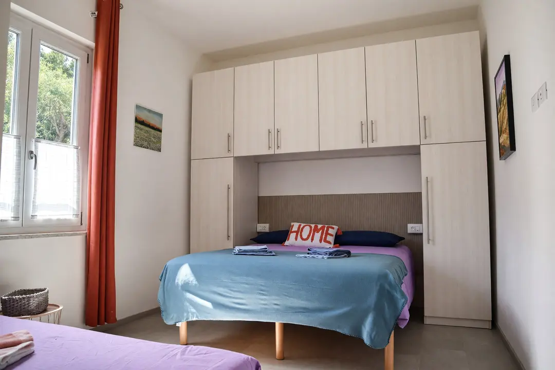 La camera dell’appartamento Lavanda: il letto matrimoniale sotto l’armadio a ponte, con la finestra e le tende rosse