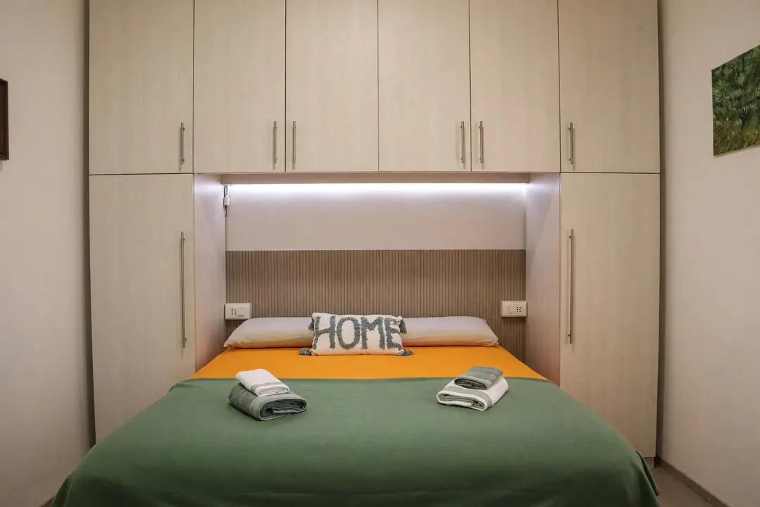 La camera dell’appartamento Ulivo: il letto con il copriletto verde e il cuscino “HOME”, sotto la luce dell’armadio a ponte