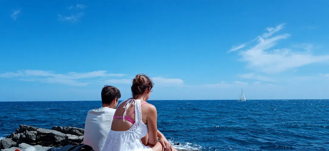 Due persone sedute sugli scogli, di spalle, guardano una barca a vela all’orizzonte
