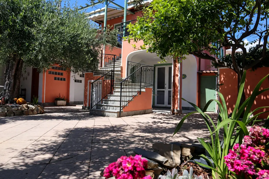 Il cortile lastricato: la scala esterna e l’arco d’ingresso della casa corallo, con i fiori rosa in primo piano