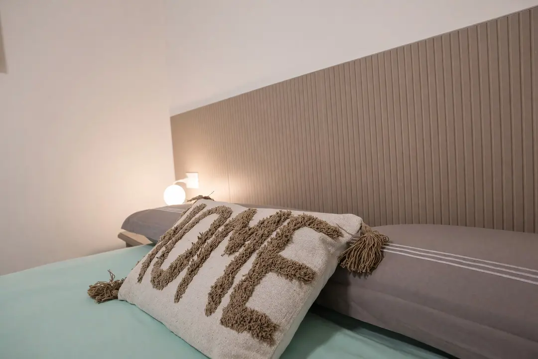 Il cuscino “HOME” sul letto dell’appartamento Basilico, con la lampada accesa