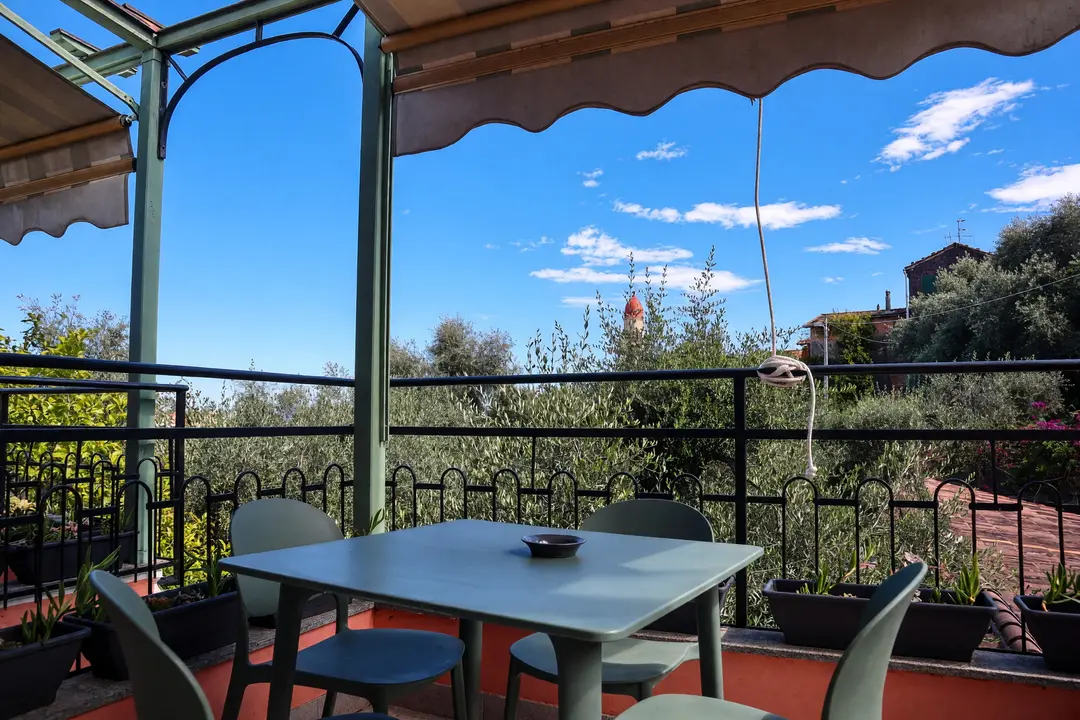Il terrazzo di un appartamento al primo piano: tavolo, sedie e tenda parasole, fra gli ulivi e il campanile